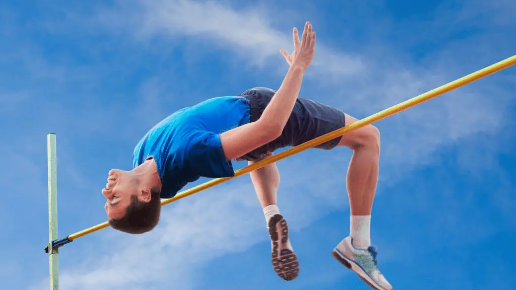 Athlete in blue shirt and shorts clears a high jump bar against a blue sky backdrop.