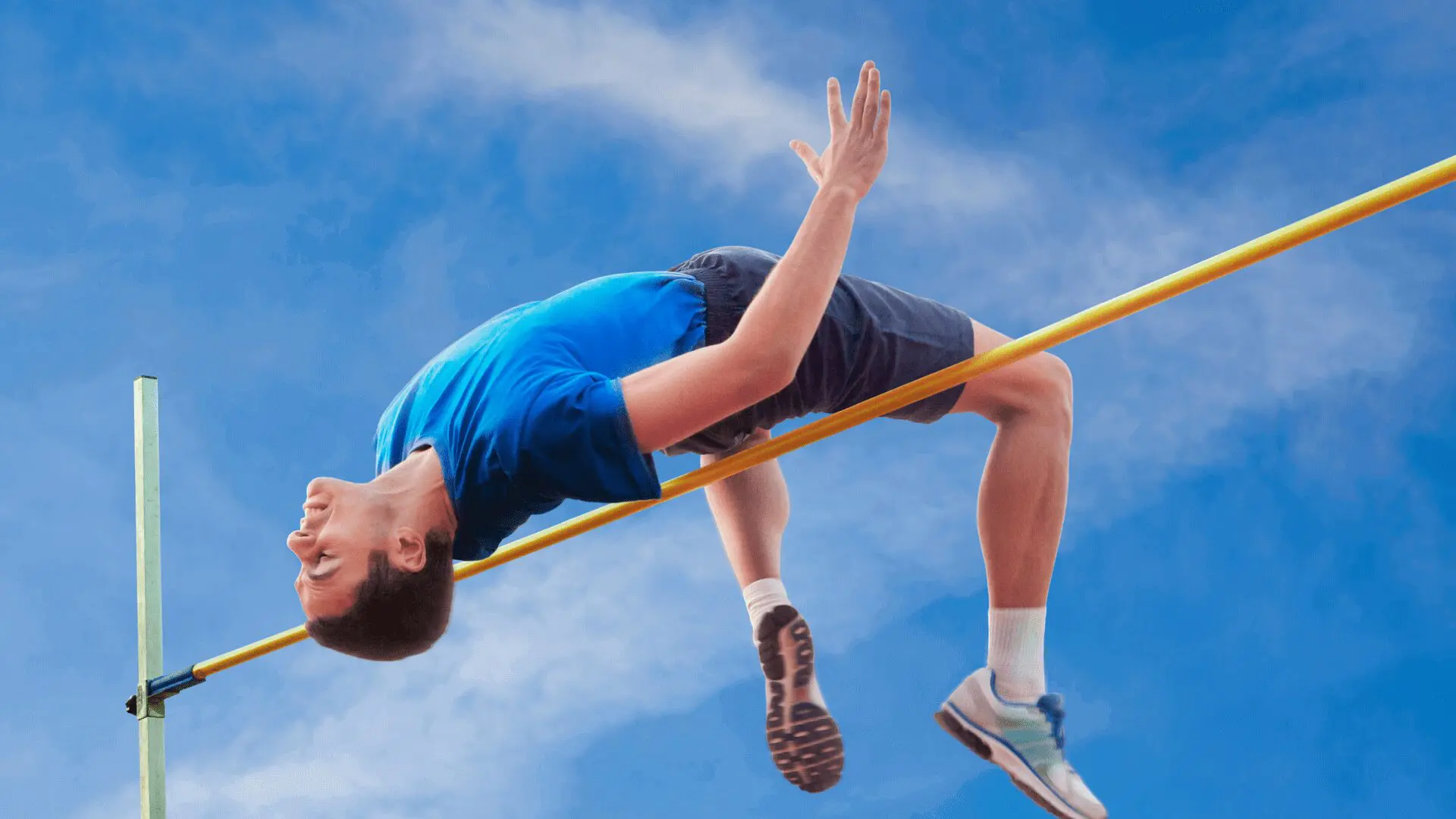 Athlete in blue shirt and shorts clears a high jump bar against a blue sky backdrop.