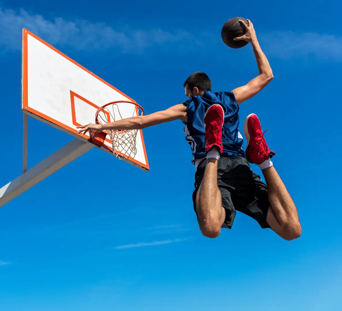A basketball player in mid-air dunking a ball into the hoop on an outdoor court under a clear blue sky.
