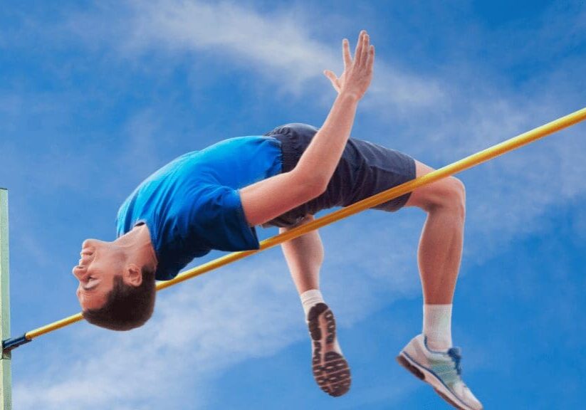 Athlete in blue shirt and shorts clears a high jump bar against a blue sky backdrop.